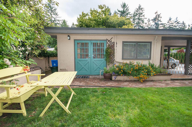 Furnished patio with vibrant yellow picnic table and green double doors, surrounded by lush greenery and a brick walkway.
