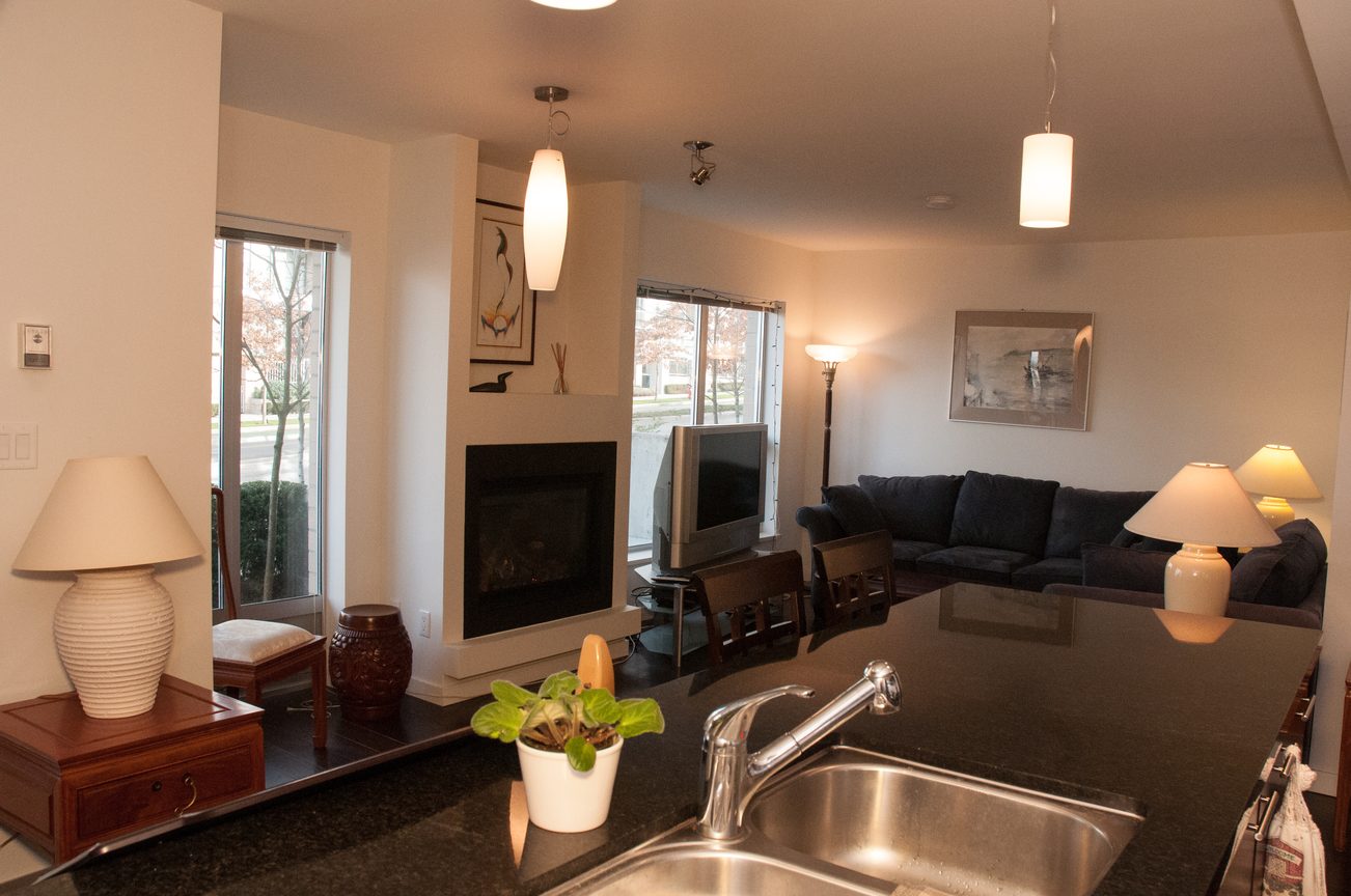 Bright living room with dark countertops and a cozy fireplace, leading to a kitchen area with a sink and potted plant.