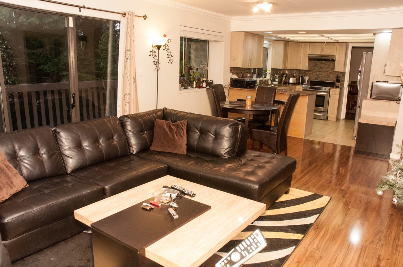 Furnished living room with hardwood floors, a sectional sofa, and a kitchen in the background. - 5550 Huckleberry Lane, North Vancouver