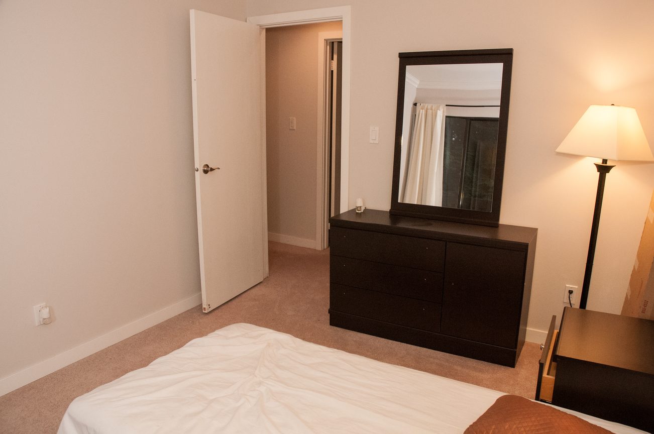 Bright bedroom with white bedding and a dark dresser, featuring a large mirror and a floor lamp. - 5550 Huckleberry Lane, North Vancouver