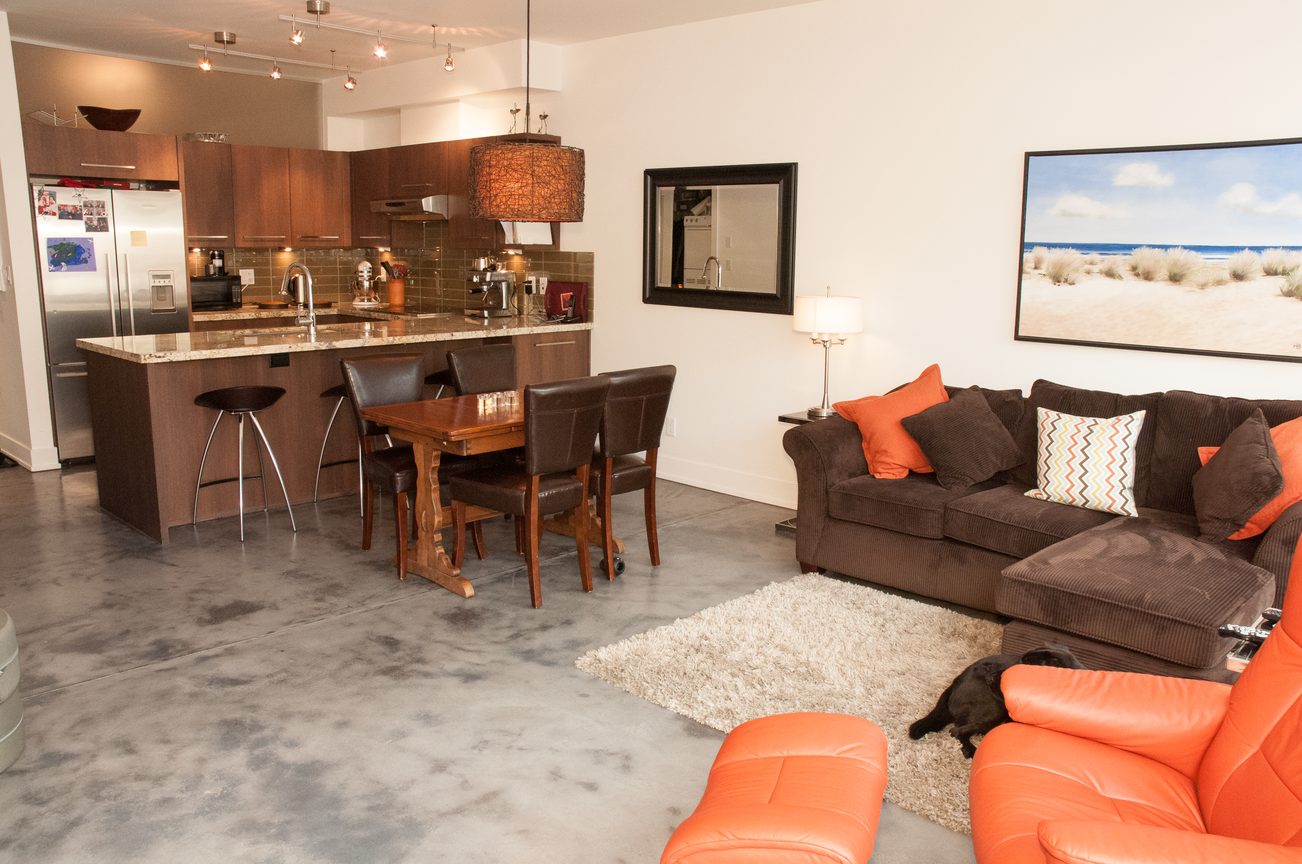 Furnished living room with orange accents and concrete floors; kitchen with granite countertops and bar stools.