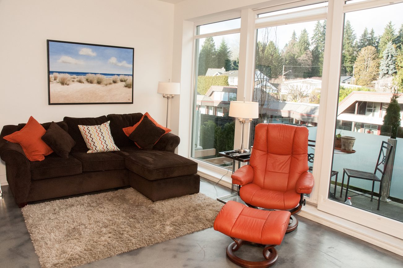 Bright living room with large windows, cozy sectional, and orange accent chair, showcasing a serene outdoor view.