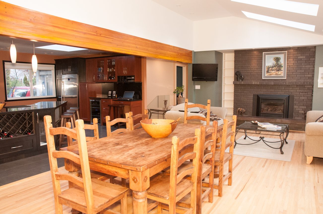 Open-concept living room with wood dining table, brick fireplace, and kitchen in the background. - 5646 Westport Rd, West Vancouver