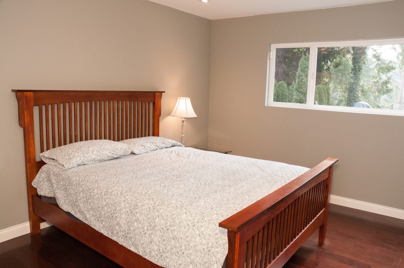 Bright bedroom with wooden bed frame, soft bedding, and a window view of greenery. - 5646 Westport Rd, West Vancouver