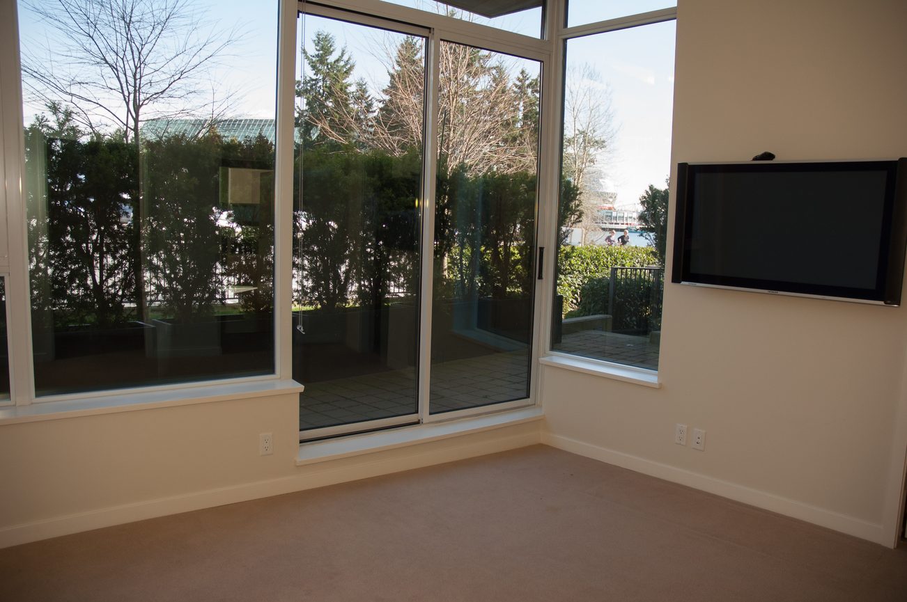 Bright living room with large windows, city view, and mounted TV. - 100-9 Smithe Mews, Vancouver Downtown