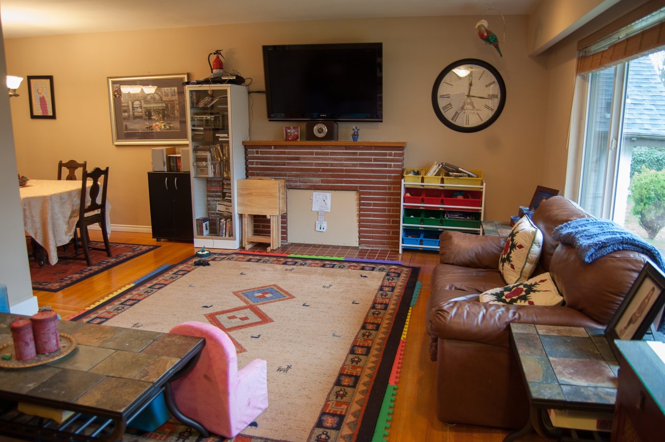 Living room with brick fireplace, colorful rugs, and a large TV mounted above. - MAIN - 451 East Keith Road, North Vancouver