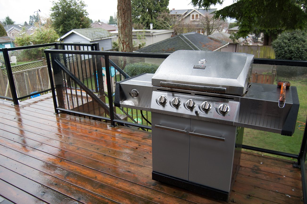 Bright deck with wet wooden flooring and a stainless steel grill, overlooking a residential neighborhood.