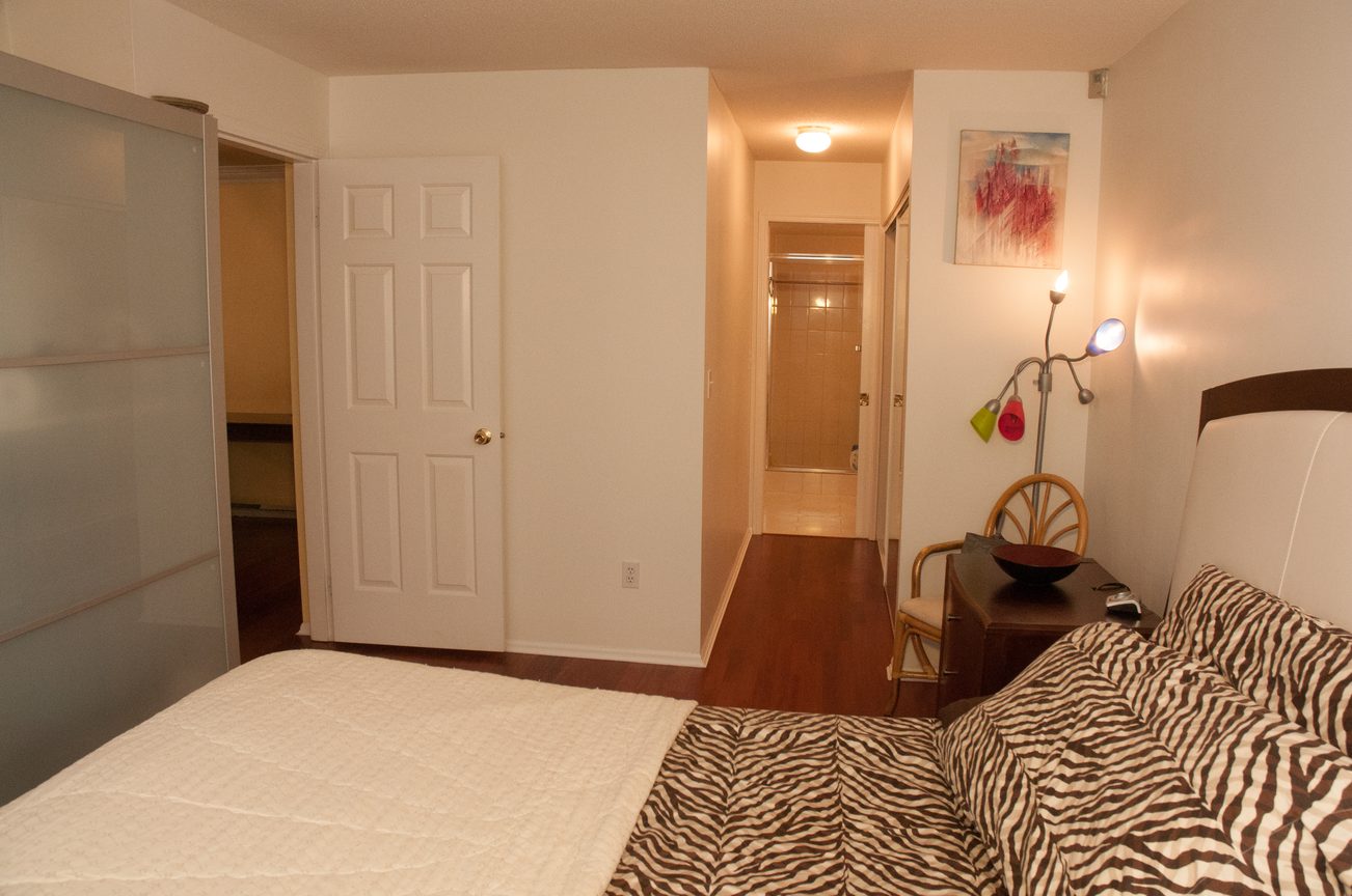 Bright bedroom with zebra print bedding and a modern lamp, leading to a hallway with a glass door. - 204 -106 West Kings Road, North Vancouver
