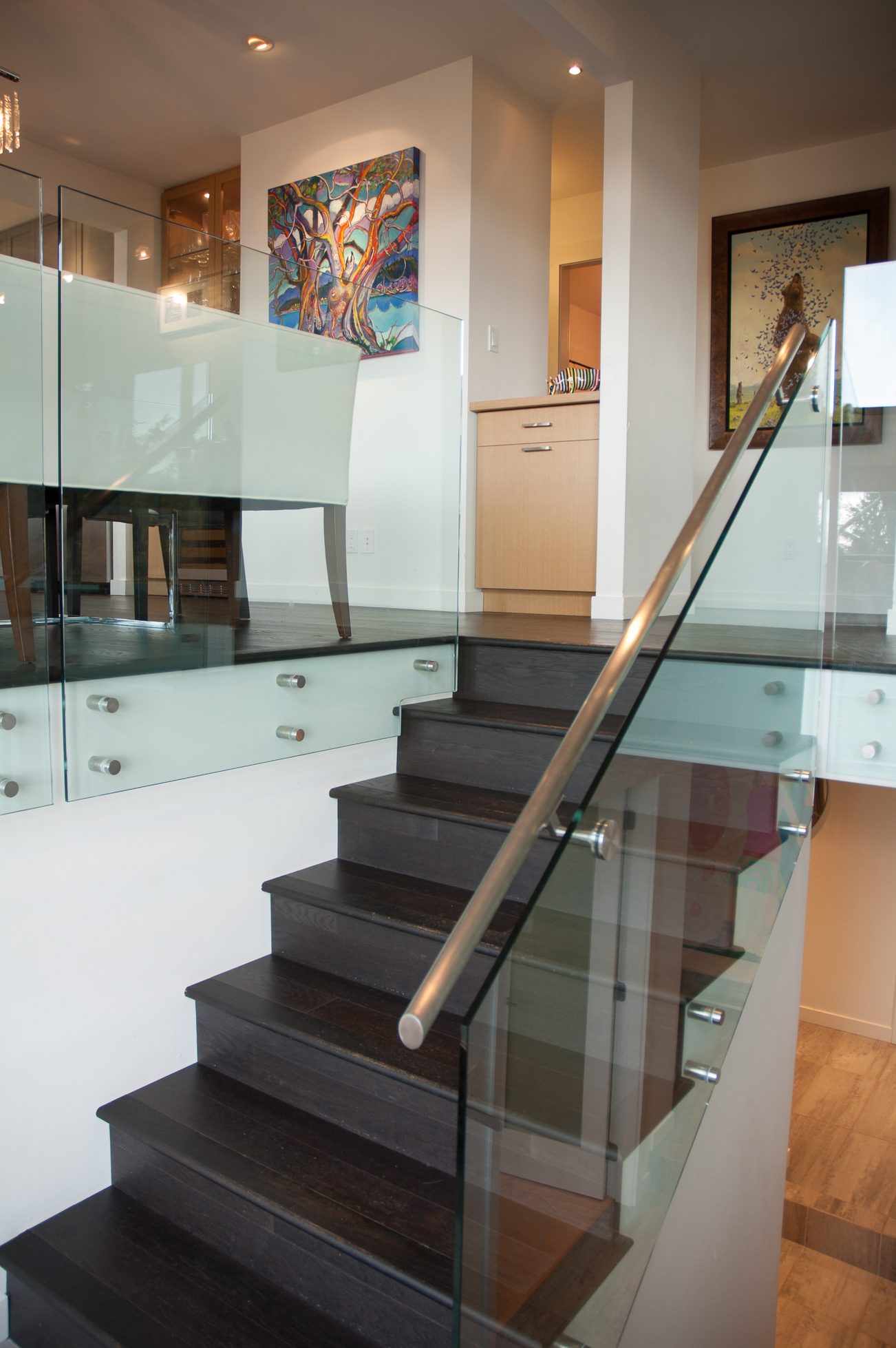 Modern staircase with glass railings leading to a well-lit dining area featuring a colorful abstract painting. - 3925 Viewridge Place, West Vancouver