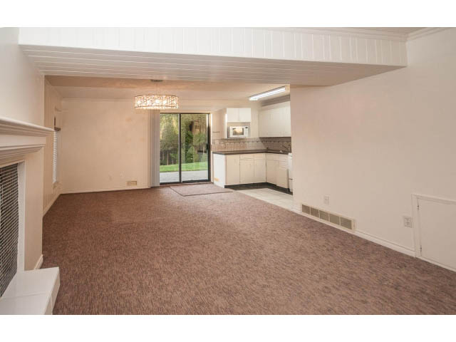 Bright living room with carpeted floors and a fireplace, leading to a kitchen with dark countertops. - Suite- 3897 St. Pauls Ave, North Vancouver
