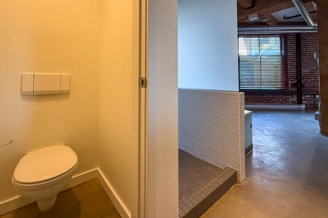 Compact bathroom with white toilet and tiled walls leading to a loft space with exposed brick and window. - 221 - 55 E. Cordova, Vancouver Downtown