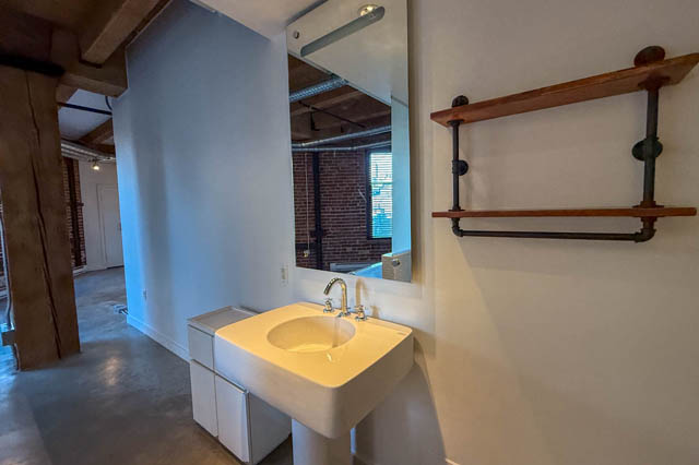 Compact bathroom with white sink, exposed brick wall, and industrial-style shelves. - 221 - 55 E. Cordova, Vancouver Downtown