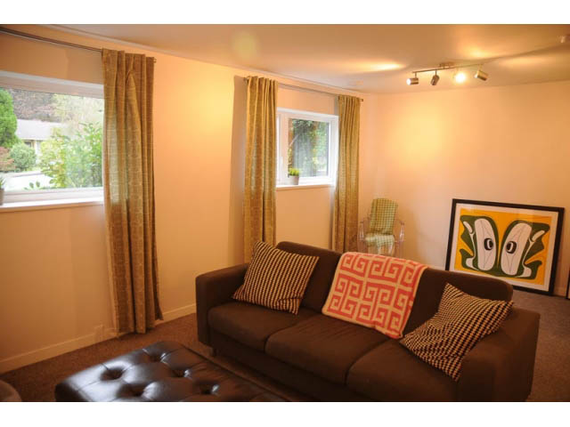 Bright living room with brown couch, colorful artwork, and natural light from windows. - 1362 East 20th St, North Vancouver