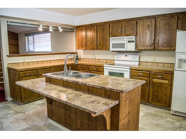 Bright kitchen with granite countertops and wooden cabinets, featuring a large island and a view of the living area.