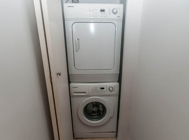 Laundry room with stacked washer and dryer, white walls, and door leading to another room. - 505 - 822 Homer Street, Vancouver Downtown
