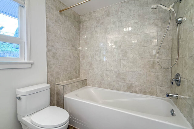 Bathroom with beige marble-look tile walls, white bathtub with handheld shower, and a window overlooking greenery. - 555 Stevens Drive, West Vancouver