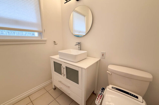 Clean bathroom with white vessel sink, glass-front vanity, round mirror, and tile floor. - 555 Stevens Drive, West Vancouver