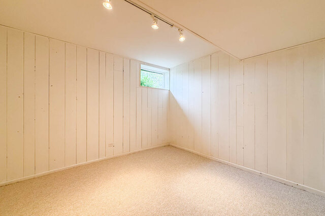 Bright room with white vertical wood paneling and a small window overlooking greenery, finished with textured carpet.