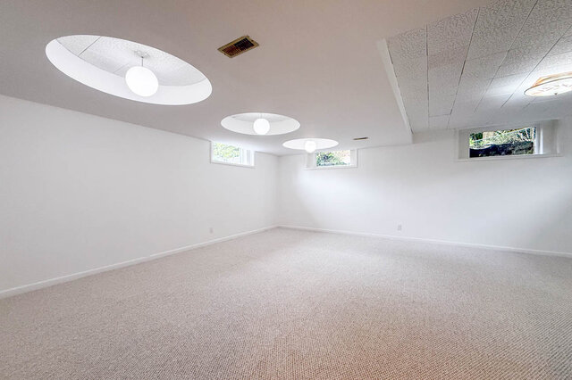 Spacious carpeted room featuring circular recessed ceiling lights with pendant globes and high windows framing garden...