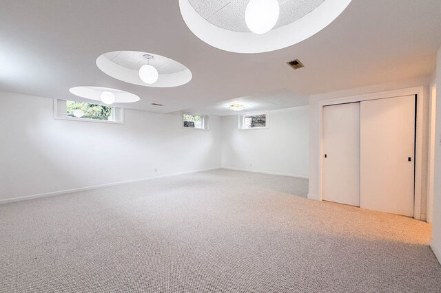 Spacious carpeted den with circular recessed pendant lights and small windows framing outdoor greenery. - 555 Stevens Drive, West Vancouver