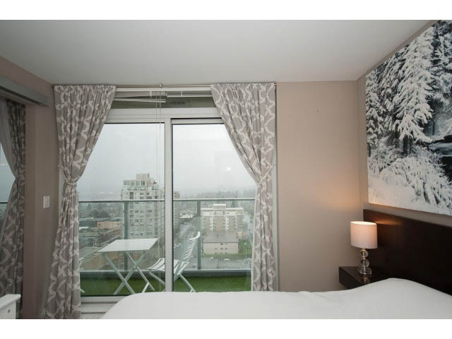 Bright bedroom with city view and modern decor, featuring a large window, white bedding, and a desk. - 1602 - 135 East 17th Street, North Vancouver