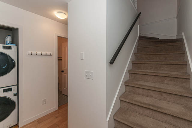 Furnished laundry room with stacked washer and dryer, hardwood floors, and a view of the staircase leading to the upper...