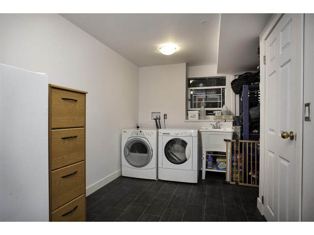 Laundry room with stacked washer and dryer, dark tile floor, white walls, and wooden cabinets. - 3243 Milton Ave., North Vancouver