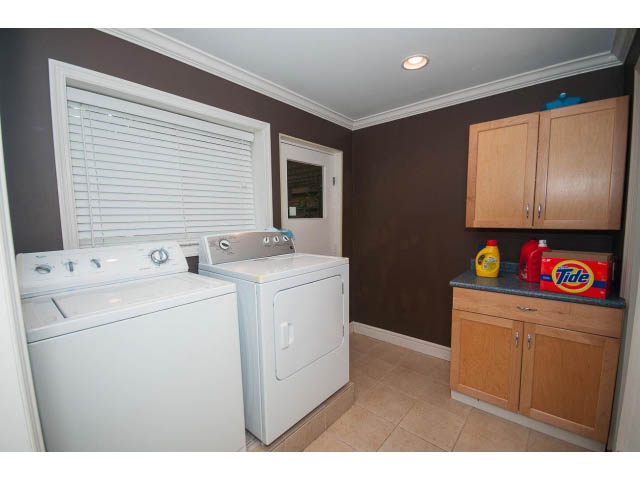 Bright laundry room with white appliances, wooden cabinets, and a window with blinds. - Upper - 1998 Cortell St., North Vancouver
