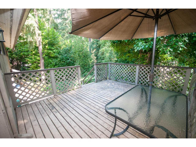 Furnished patio with wooden deck, glass table, and umbrella, surrounded by lush greenery. - Upper - 1998 Cortell St., North Vancouver