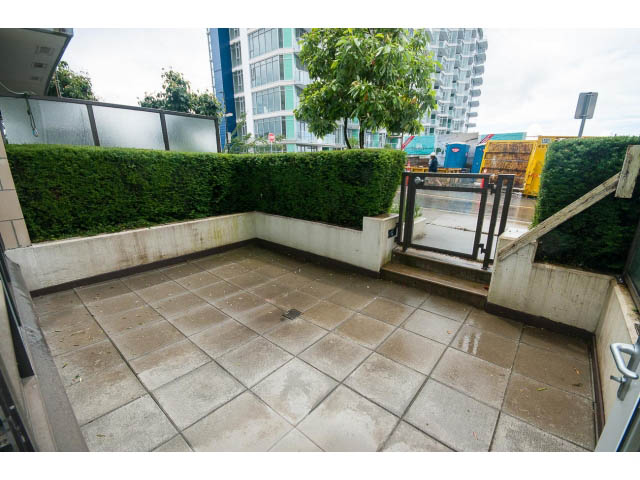 Bright patio with tiled flooring and greenery, leading to a modern building in the background. - 203 - 162 Victory Ship Way, North Vancouver