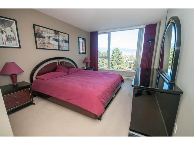 Furnished bedroom with a large window offering a city view, red bedding, and a sleek dresser. - 603 - 1425 West 6th Ave, Vancouver West Side