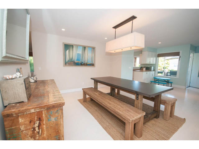 Bright dining area with wooden benches and a modern chandelier, leading to a kitchen with light blue walls and a city view.