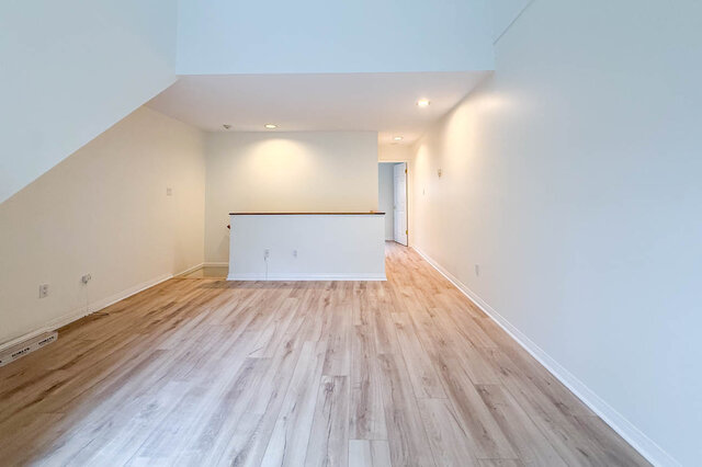 Long living room with hardwood floors and pot lights, half wall and a hallway leading to the bedroom and bathroom.