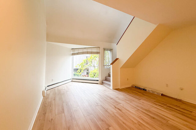 Living room with sloped ceiling, hardwood floors, corner picture window onto green treetops and the stairs up to the loft.