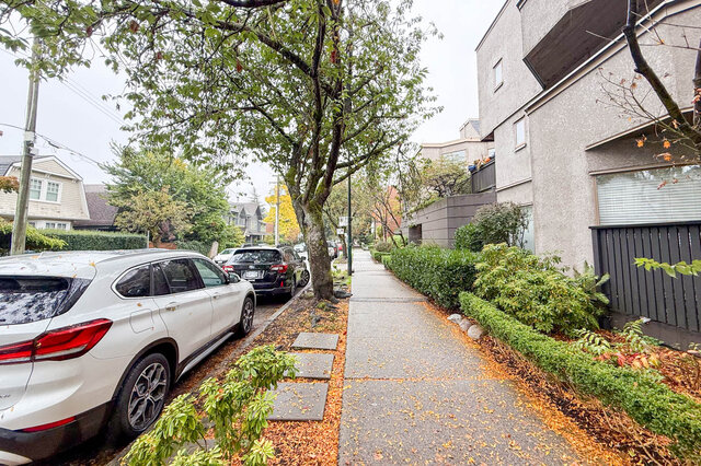Leafy residential street beside the townhouses: wide sidewalk, street trees, trimmed hedges and parked cars.