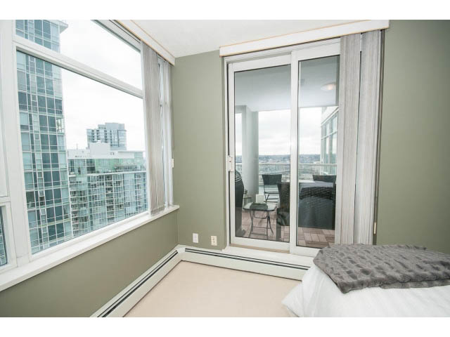 Bright bedroom with large windows offering a city view, furnished with a bed and a sliding glass door leading to a balcony.