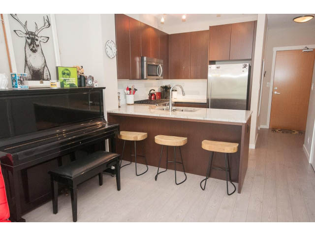 Bright kitchen with wooden cabinets, black piano, and hardwood floors. - 203 - 135 West 2nd Street, North Vancouver