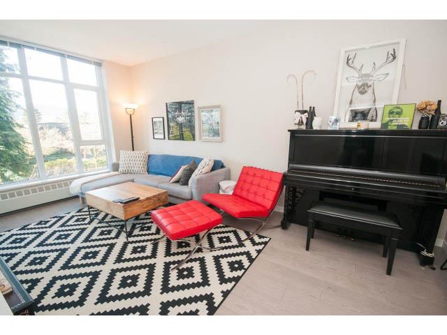 Furnished living room with red ottoman, black piano, and geometric rug. - 203 - 135 West 2nd Street, North Vancouver
