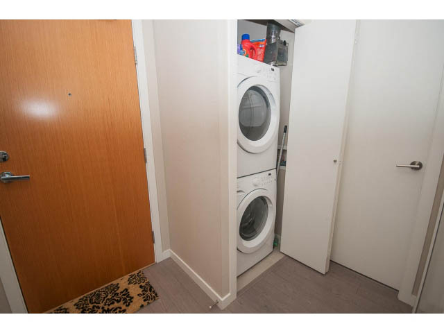 Laundry room with stacked washer and dryer, wooden door, and small rug. - 203 - 135 West 2nd Street, North Vancouver