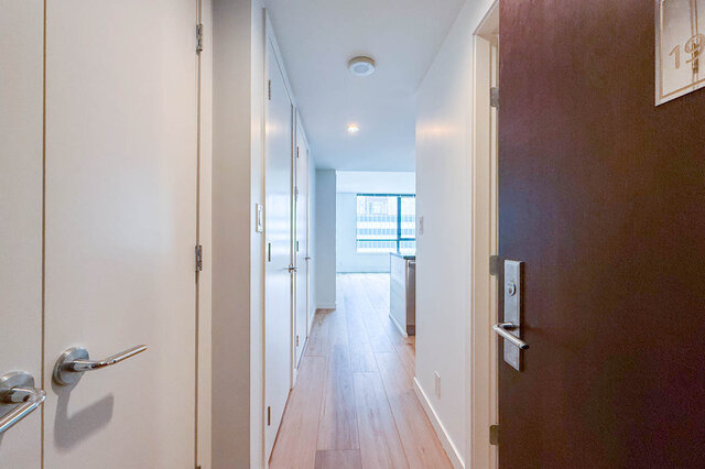 Entry hallway with light hardwood floors, white walls, and a dark wood door opening toward a bright windowed kitchen.