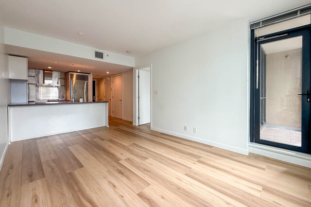 Living room with light hardwood floors and an open kitchen with stainless steel appliances and a balcony door.