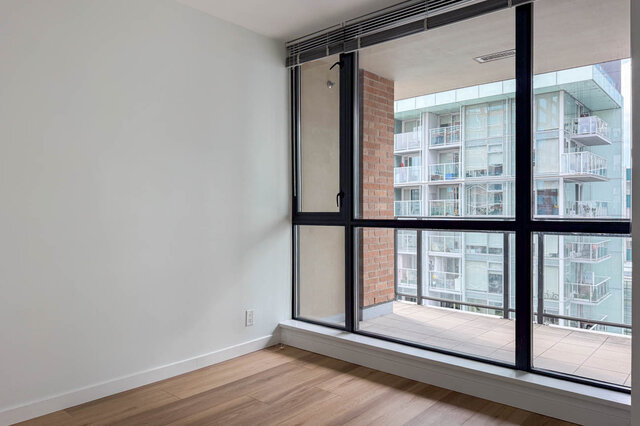 Empty living room with light hardwood floors and a large black-framed window overlooking a neighboring condo building.