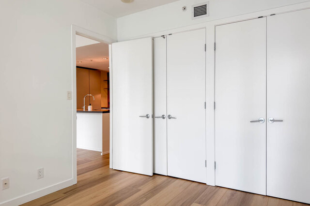 Bright bedroom with white built-in closet doors and light hardwood floors, opening to a kitchen with dark countertops.