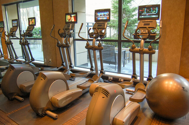 Fitness room with elliptical trainers, wall-mounted TV screens, and floor-to-ceiling windows overlooking a patio.