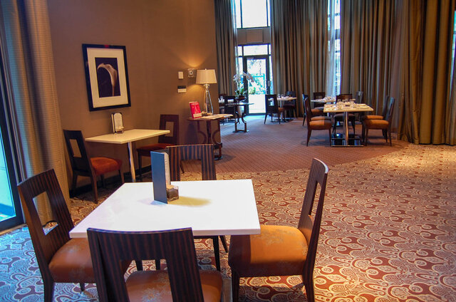 Dining area with white-topped tables, amber-upholstered chairs, and patterned carpet beneath tall draped windows.