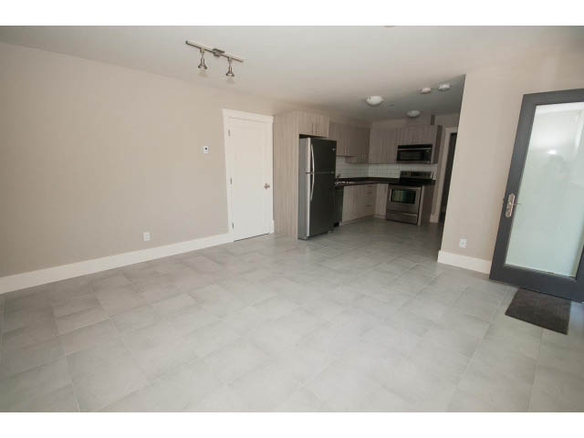 Spacious living room with tiled floor and kitchen area featuring stainless steel appliances and light wood cabinets.