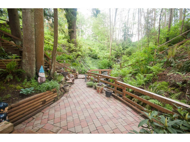 Spacious outdoor patio with wooden railings and brick flooring, surrounded by lush greenery. - Suite - 2985 Dollarton Hwy, North Vancouver