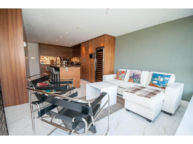 Furnished living room with white leather couches, modern bar stools, and a sleek kitchen in the background.
