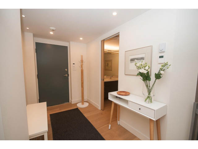 Bright hallway with white walls, wooden floors, and a black rug leading to a door and a small desk area.