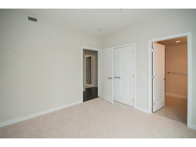 Bright living room with white walls and carpeted floor, leading to a den and hallway. - 303 - 3825 Cate's Landing, North Vancouver